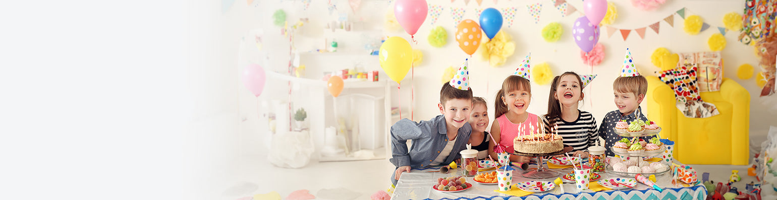 Fünf Kinder mit Partyhüten stehen vor einem Tisch mit Kuchen, Süßigkeiten und Muffins, im Hintergrund bunt geschmückt.