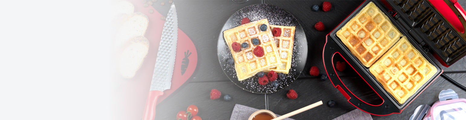 Ein gefülltes Waffeleisen und fertige Waffeln mit Himbeeren und Blaubeeren auf einem schwarzen Tisch.