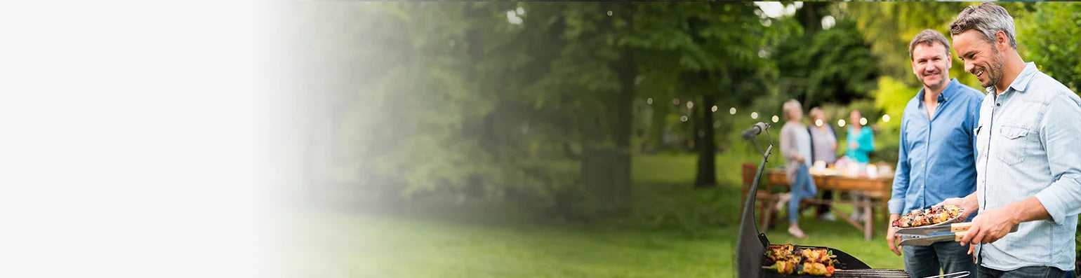 Zwei Männer stehen an einem Grill, im Hintergrund Frauen an einem gedeckten Tisch in einem Garten mit vielen Bäumen.