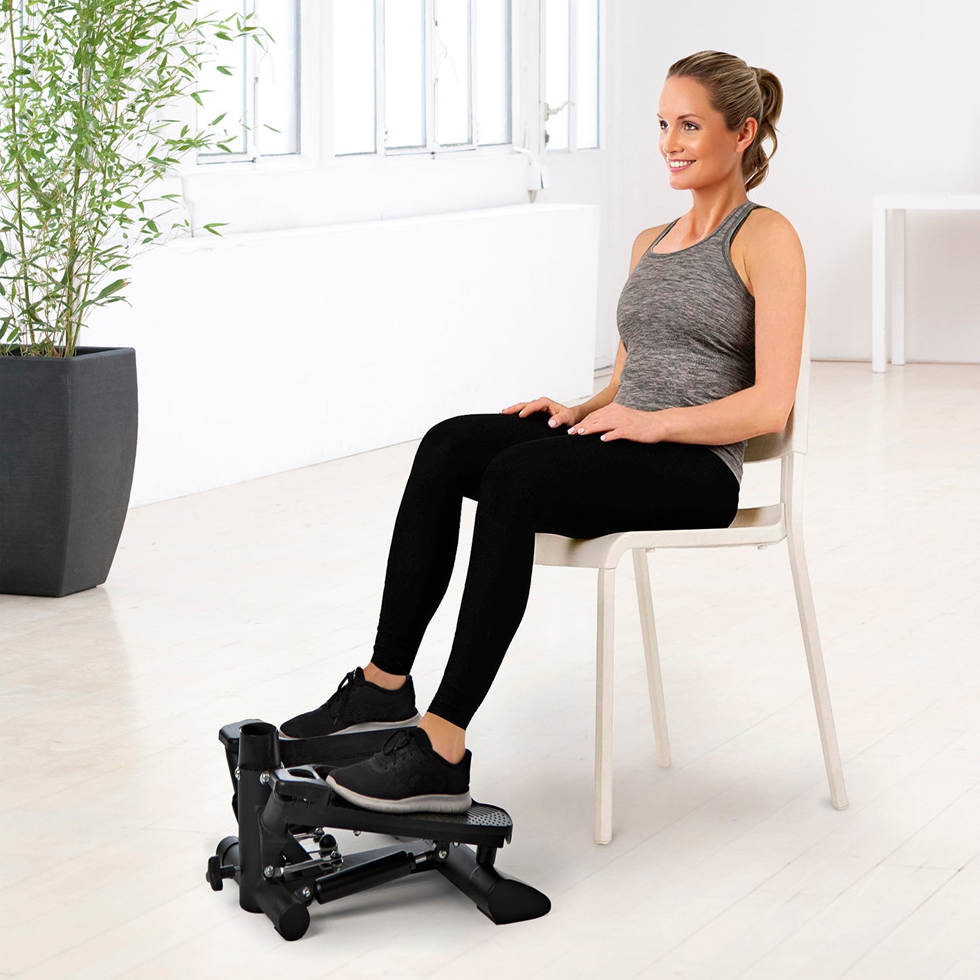 Frau trainiert sitzend mit Fitnesstrainer Swing Stepper mit 2 Expanderbändern im hellen Raum für Beine und Arme.