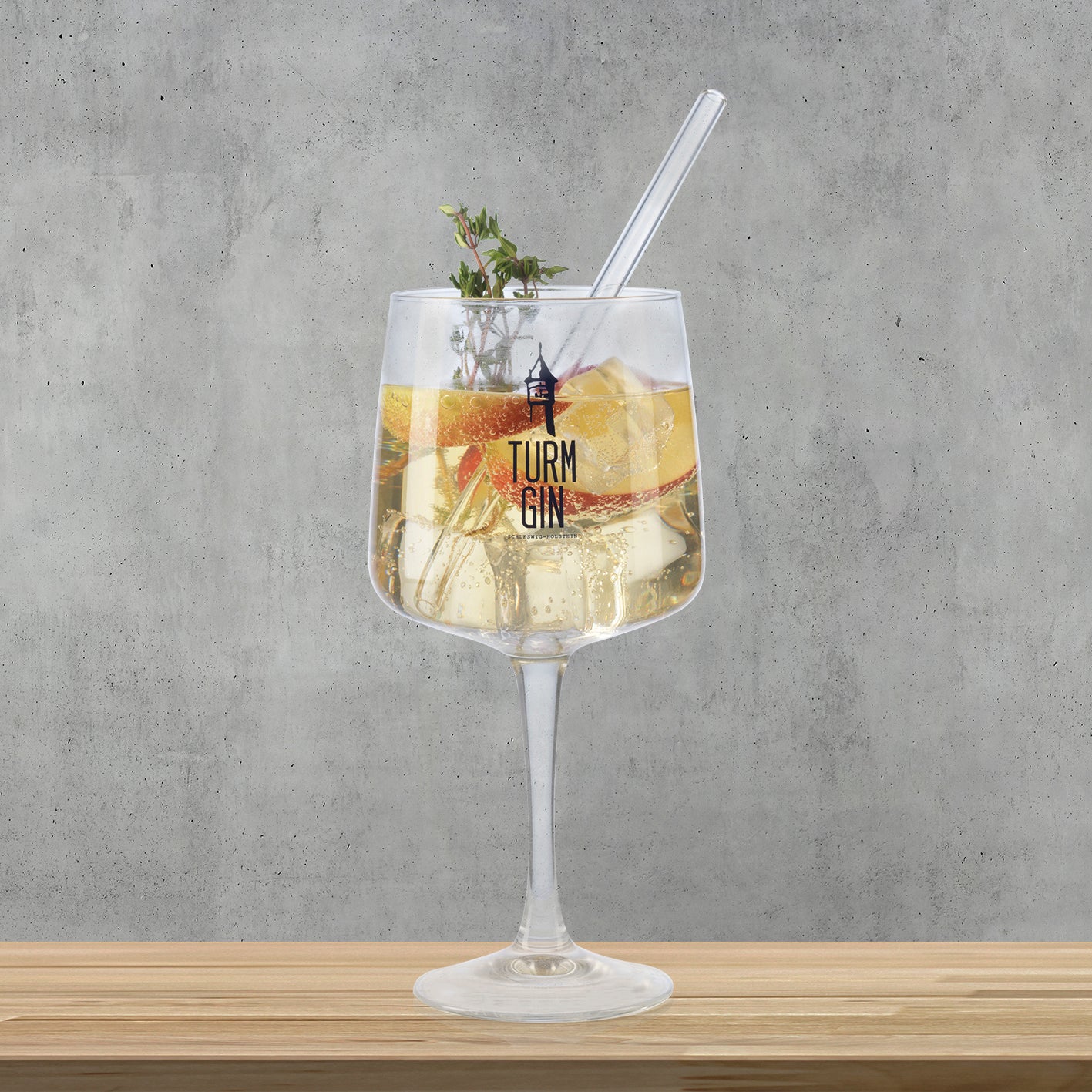 Copa Glas mit Logo und Schriftzug - 720 ml - 2er-Set, stilvolles TURM GIN Glas mit Getränk, Eis und Garnitur auf Holz vor grauer Wand.