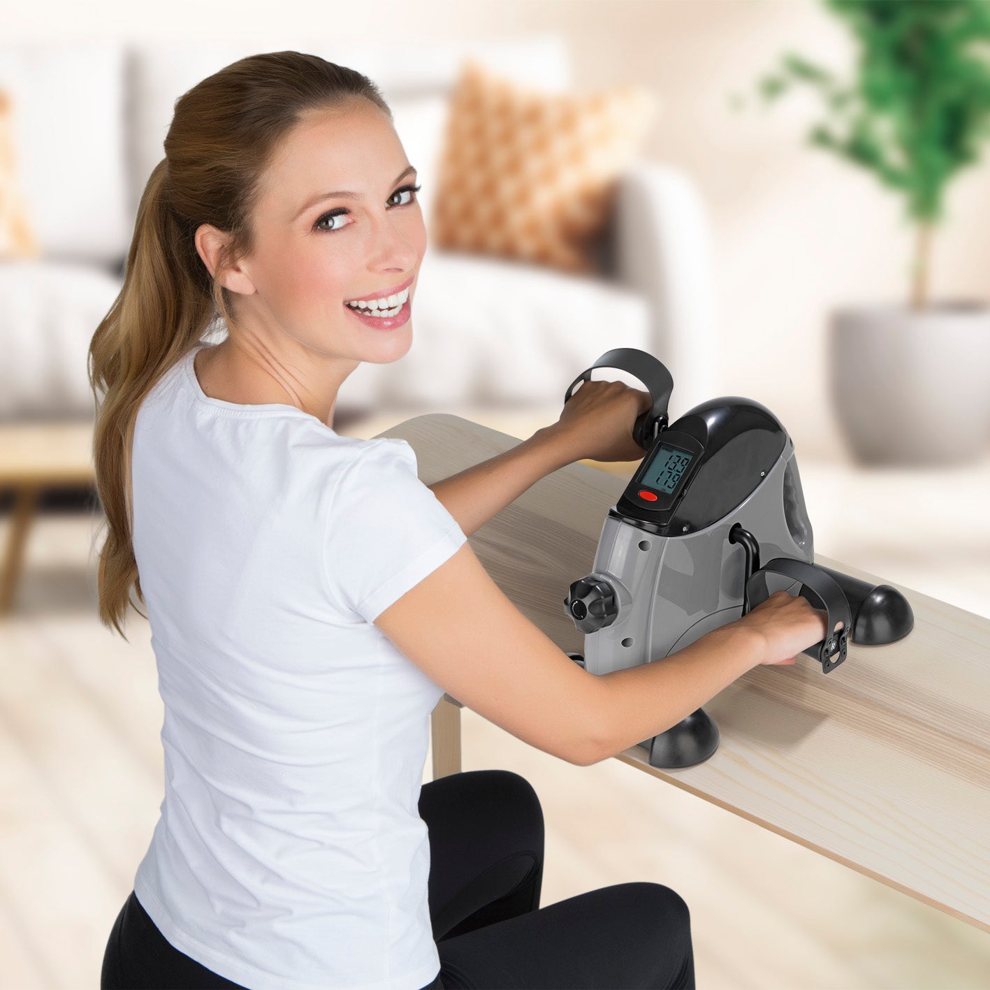 Frau trainiert Arm- und Beinmuskulatur mit Mini-Trainer mit Metallschwungrad - grau/schwarz am Tisch mit Trainingscomputer.