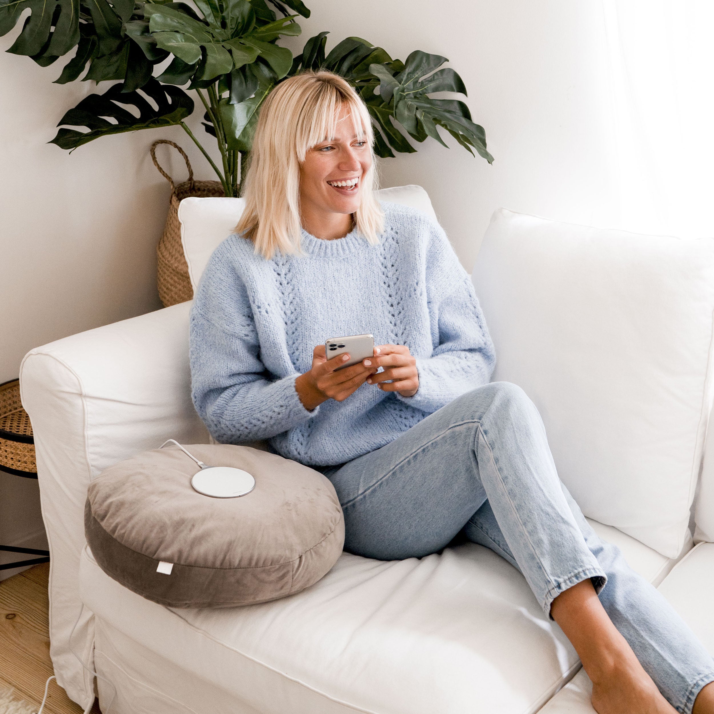 Frau entspannt auf Sofa mit Smartphone und silbernem Wireless Ladegerät Aura lite 15W neben ihr auf grauem Kissen.
