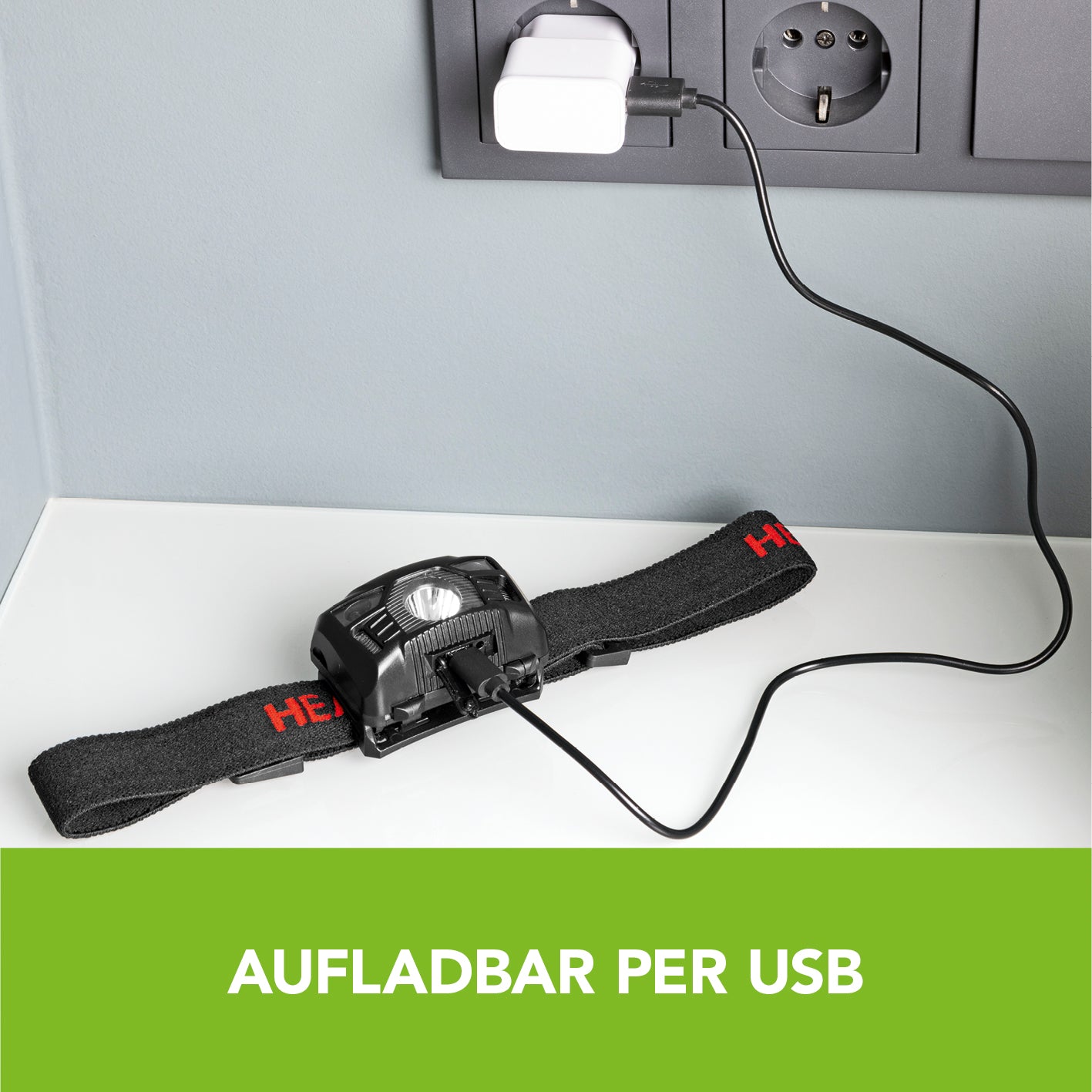 Stirnlampe schwarz/rot mit USB-Ladekabel auf weißem Tisch vor Steckdose, wiederaufladbar und mit verstellbarem Kopfband