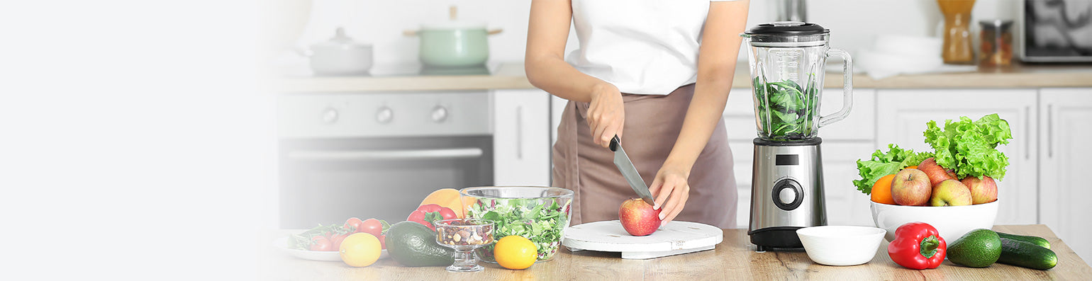 Eine Frau in einer Küche schneidet einen Apfel, neben ihr diverses Obst und Gemüse und einem Mixer. 