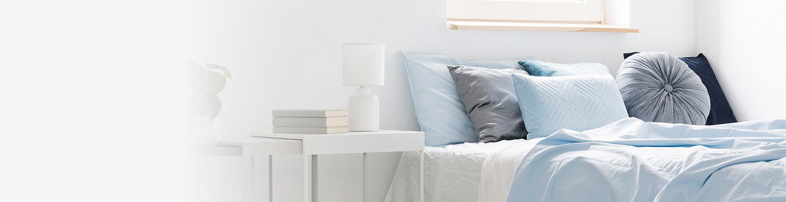 Ein Bett mit diversen hellblauen und grauen Kissen, daneben ein weißer Nachttisch.
