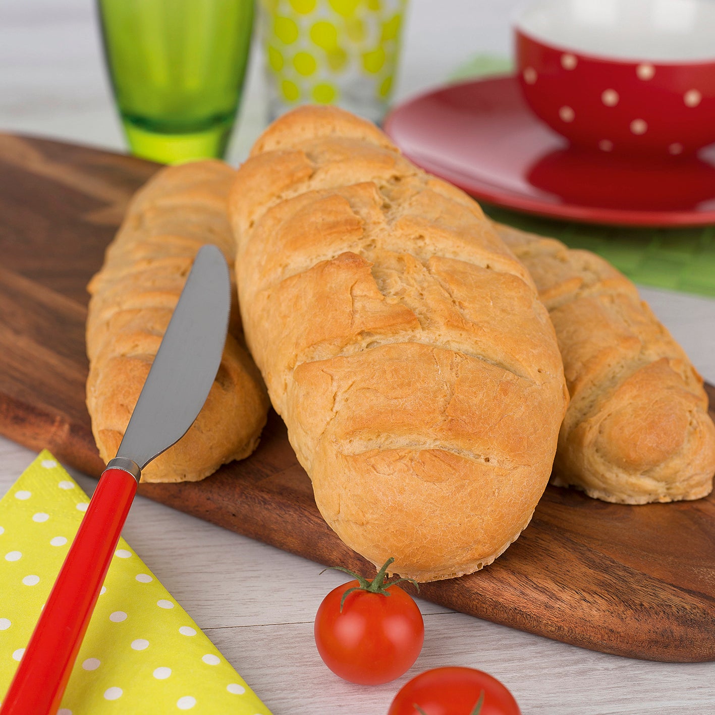 Rezeptbuch inkl. Messbecher-Set - Band 4 - Kleine Gerichte ganz groß! zeigt frische Brote auf Holzbrett mit Tomaten und Messer.
