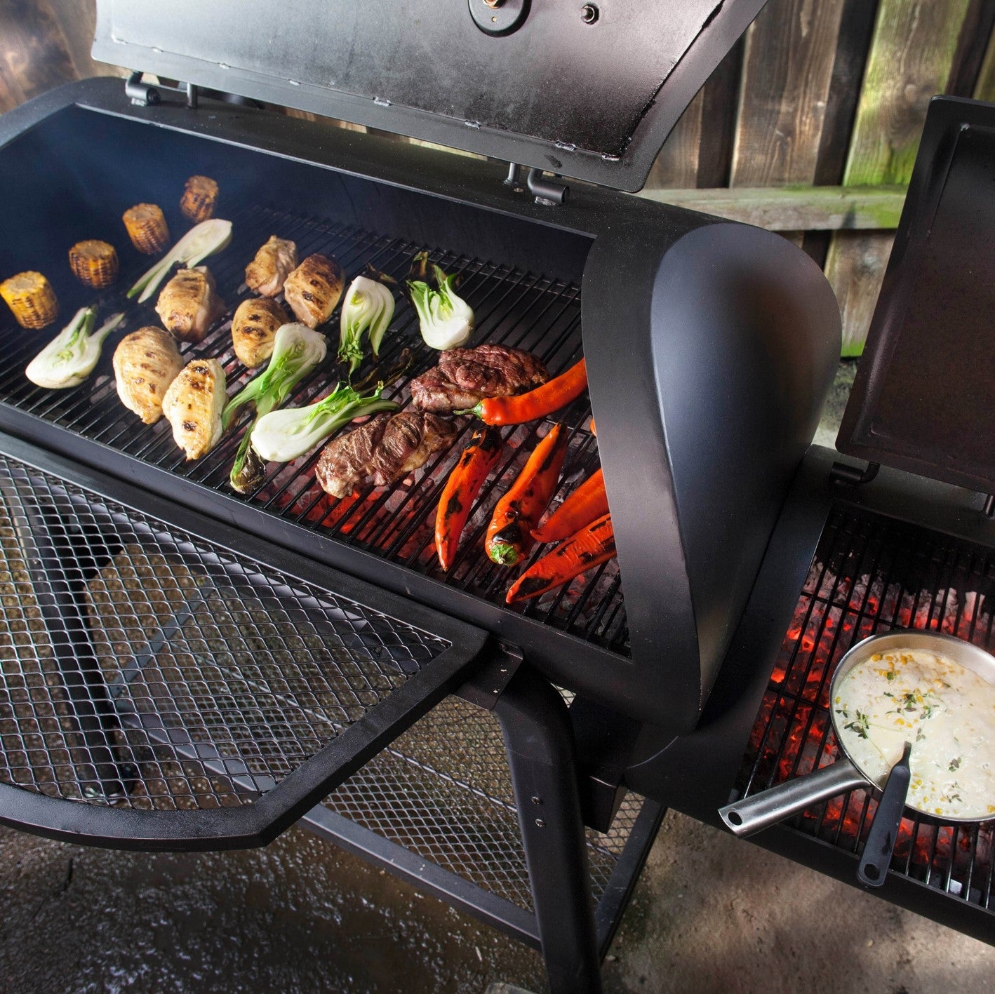 Smoker Vinson 500 mit offenem Deckel, gegrilltem Gemüse und Fleisch, Edelstahlgriffen und 4-in-1 Funktion für direktes und indirektes Grillen.