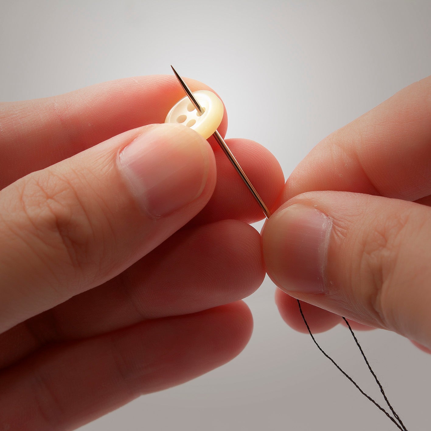 Vergrößerungsbrille LED - schwarz zeigt nähende Hände, die eine Nadel mit Faden durch einen Knopf fädeln.