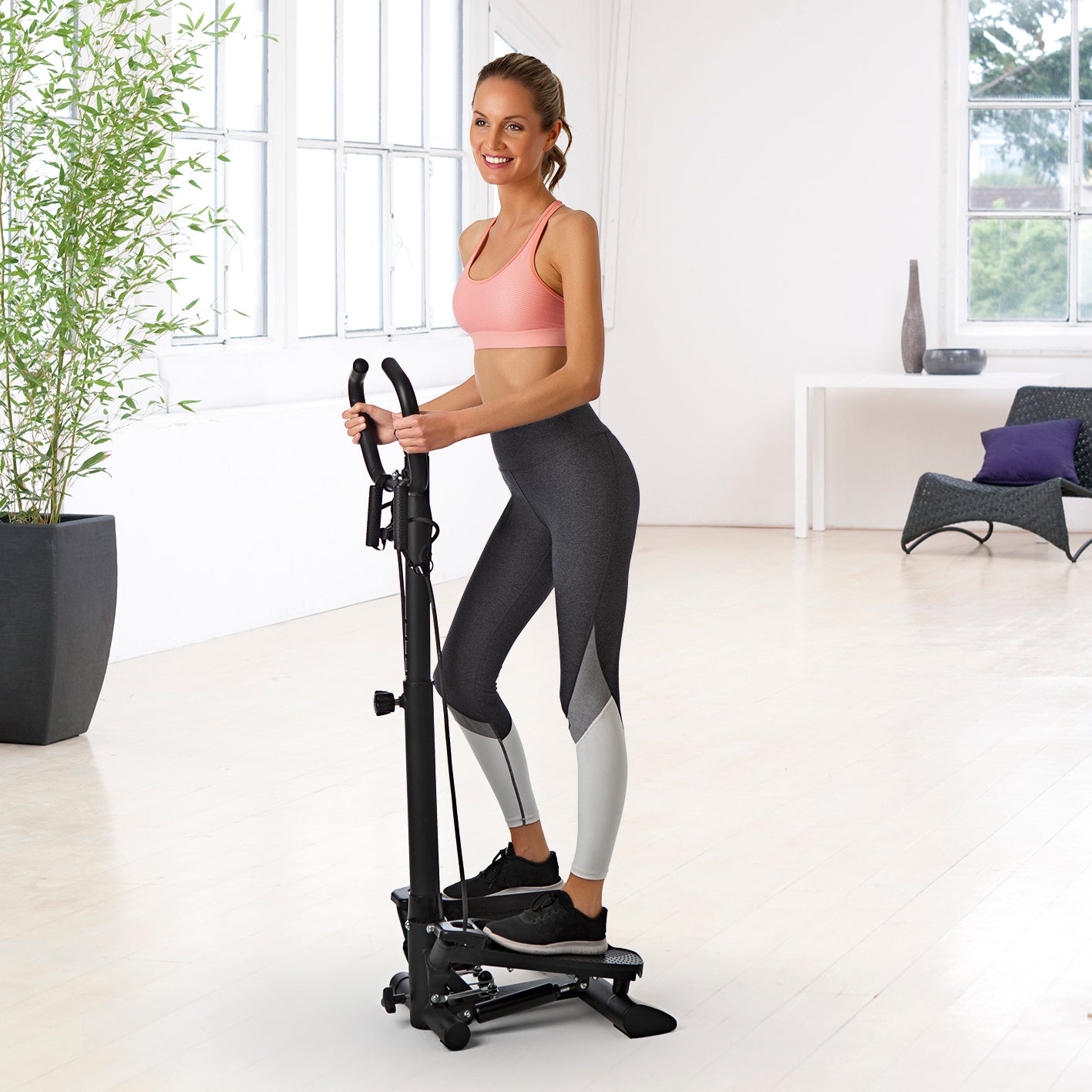 Frau trainiert mit Fitnesstrainer Swing Stepper mit 2 Expanderbändern in hellem Raum mit großem Fenster und Pflanze.