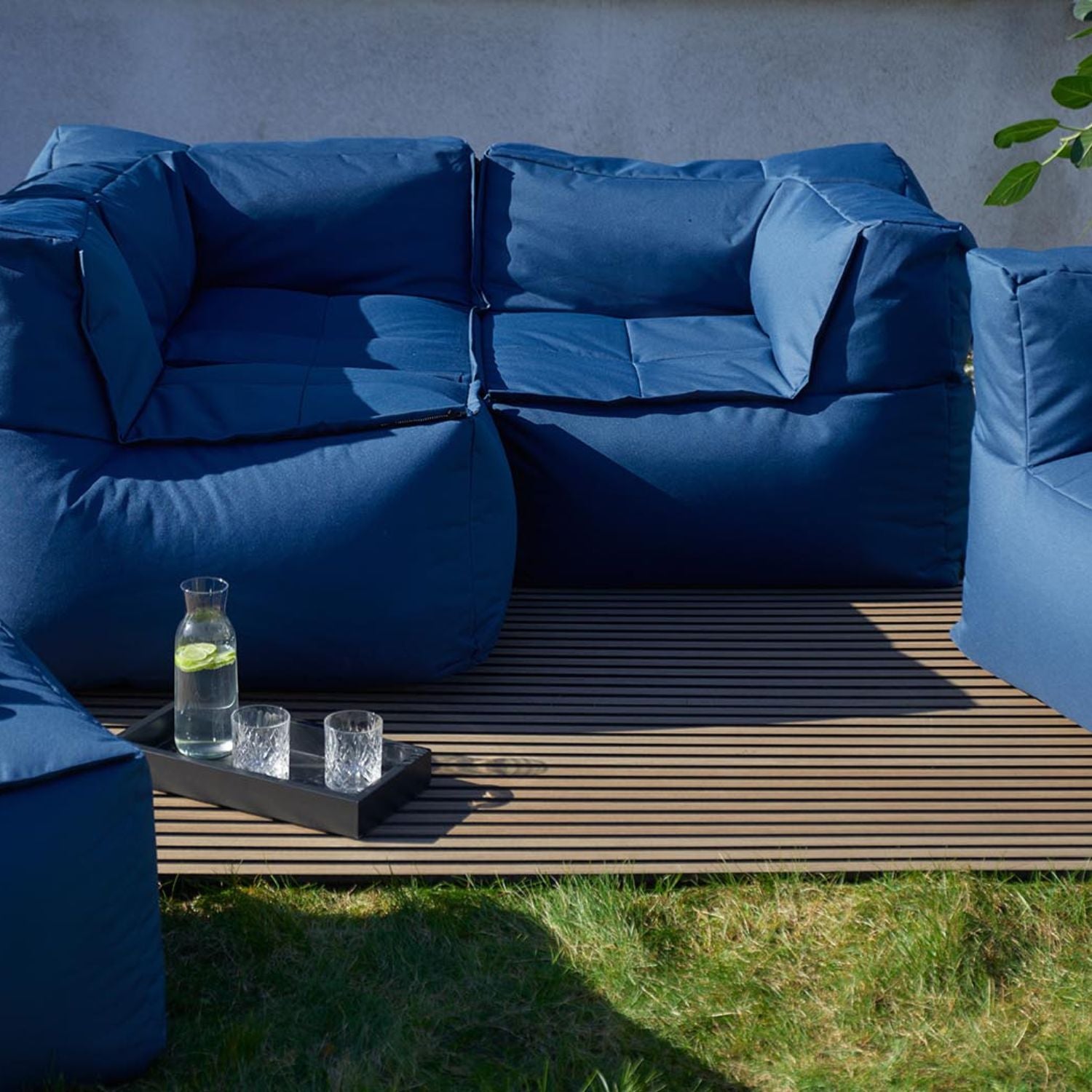 Moderne LUMALAND Outdoor-Lounge mit blauen Polstersesseln auf Holzdeck und Tablett mit Karaffe und Gläsern auf grünem Rasen.